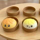 Orange and pale yellow squishy toys sit in open brown round containers on a wooden table, with matching lids behind them.