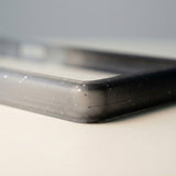 Close-up of a translucent black-gray slab bumper corner shows pale speckles and a recessed edge on a light surface.