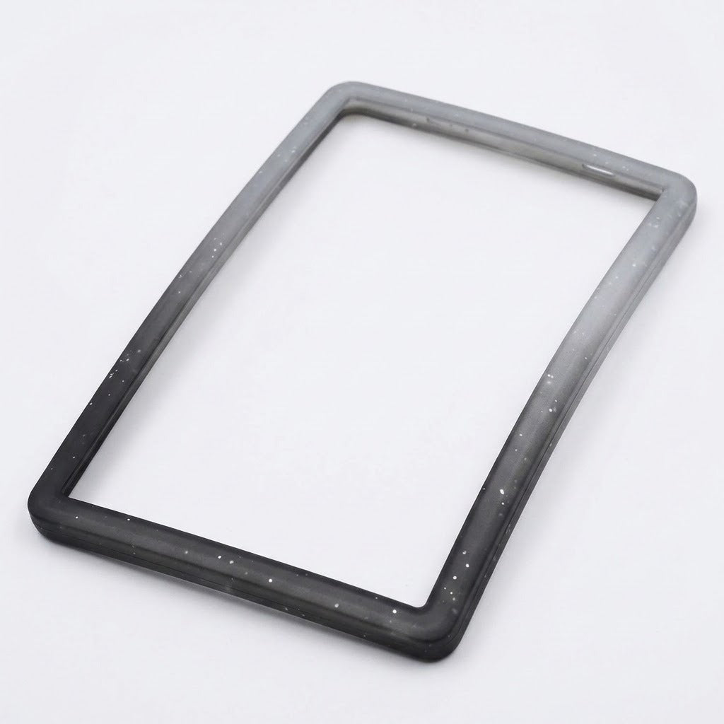 A speckled gray-to-black silicone TPU slab bumper lies diagonally on a white background, showing its open center and rounded corners.