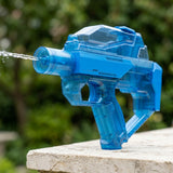 A translucent blue electric water gun stands on a pale stone surface, spraying water against a blurred green garden background.