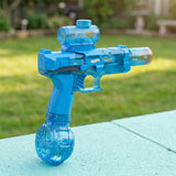 A translucent blue electric water gun with cylindrical top tank and round base stands on a pale blue surface outdoors.