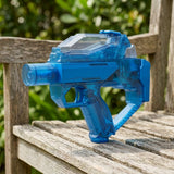 A translucent blue electric water gun with a clear top tank rests on a weathered wooden bench against blurred greenery.