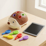 Brown and cream hedgehog sensory toy with colored openings sits on a light wooden table beside five colorful pegs and a tablet.