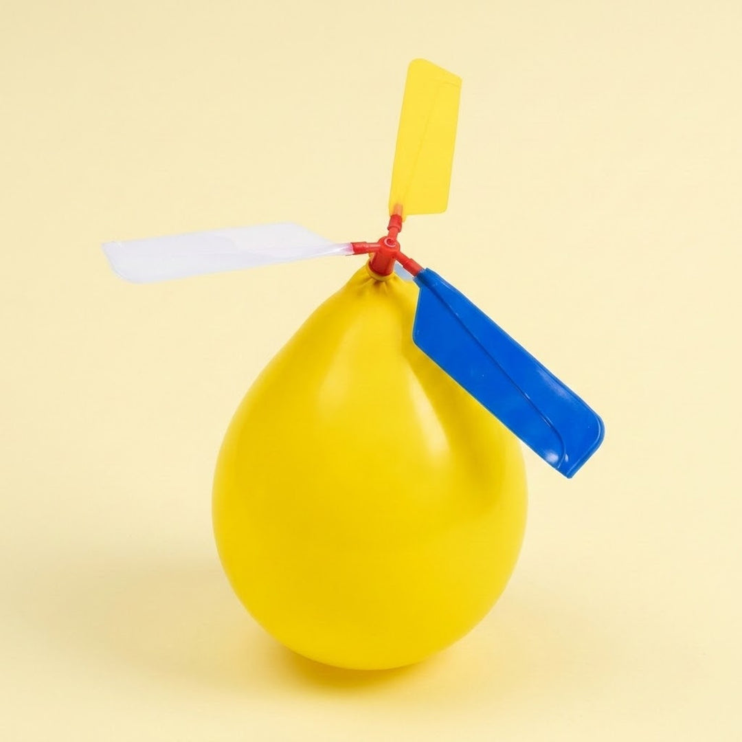 A balloon helicopter with an inflated yellow balloon and blue, white, and yellow plastic propeller blades on a red connector against a pale yellow background.