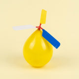 An assembled balloon helicopter set has an inflated yellow balloon with yellow, white and blue plastic propeller blades against a pale yellow background.