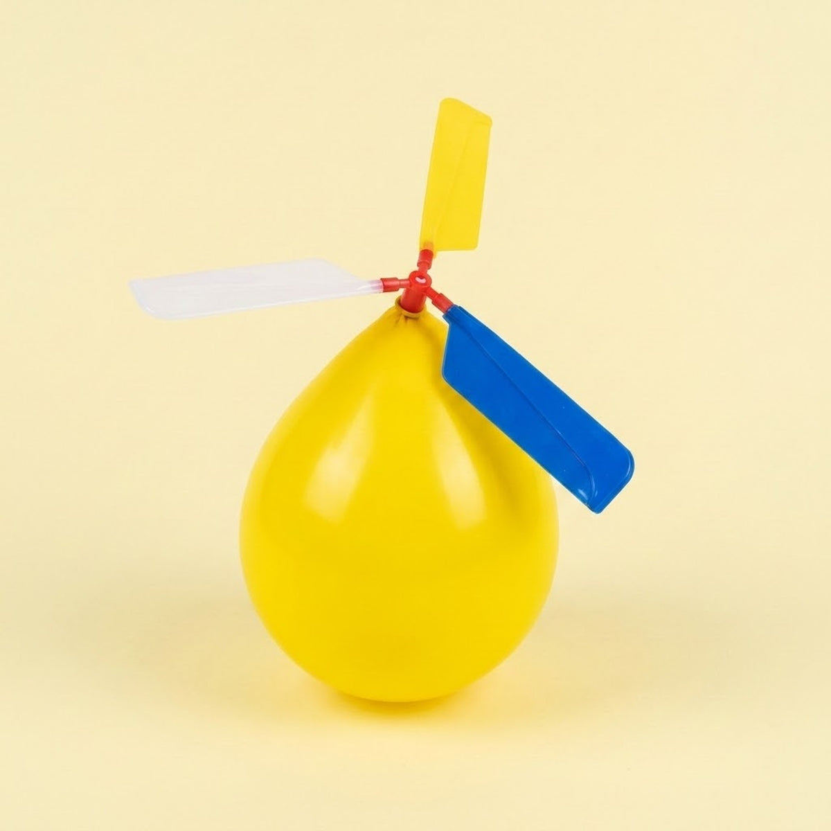 An assembled balloon helicopter set has an inflated yellow balloon with yellow, white and blue plastic propeller blades against a pale yellow background.