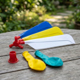 Balloon helicopter set components include colourful plastic propeller blades and deflated balloons on a weathered wooden table against blurred garden greenery.