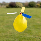 A yellow balloon-powered toy helicopter with a white and blue propeller, photographed outdoors against a blurred green grassy background.