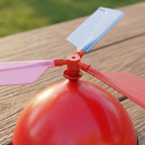 A balloon helicopter set close-up shows a red balloon beneath light blue, pink and coral plastic propeller blades on an outdoor wooden table.