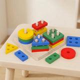 A colorful wooden shape-sorting toy set arranged on a small light wood table. The set includes round and geometric blocks in yellow, blue, green, red, and white pegs, with loose matching pieces around them.