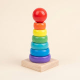 Wooden educational toy with rainbow stacking rings and a red ball on a square natural wood base against a beige background.