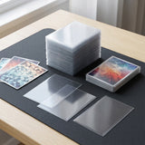 A stack of clear plastic card sleeves and several individual transparent sleeves arranged on a dark mat on a light wood table, with some colorful card backings visible.