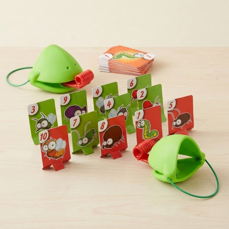 Frog tongue party game with two green masks, red tongues, upright numbered insect targets, and stacked cards on a pale tabletop.