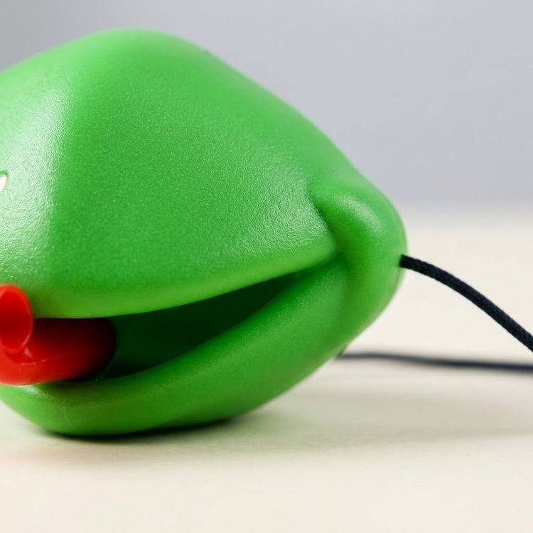 Frog tongue party game mask shown close up with a textured green surface, red tongue, and black securing cord against a pale background.