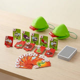 Frog tongue party game with two green masks, numbered insect targets, fanned orange cards, and a stacked deck on a pale wooden table.