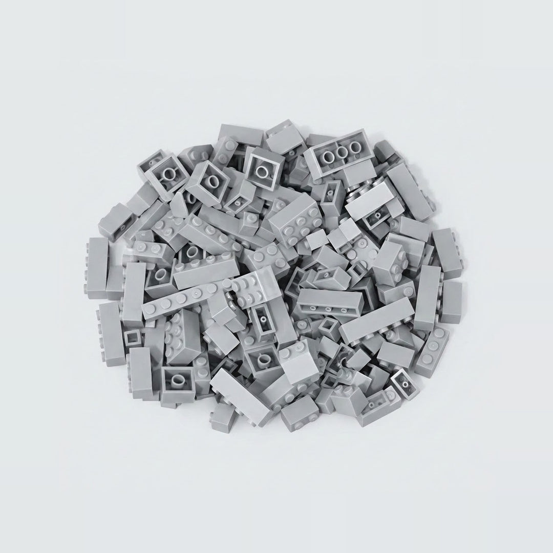 A compact pile of light gray building bricks rests on a pale background, with square and rectangular pieces facing different directions.