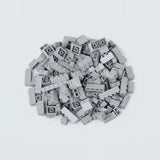 A compact pile of light gray building bricks rests on a pale background, with square and rectangular pieces facing different directions.