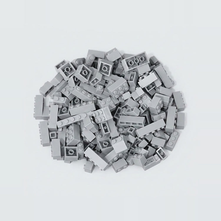 A compact pile of light gray building bricks rests on a pale background, with square and rectangular pieces facing different directions.