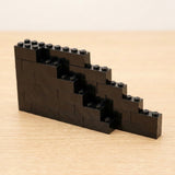 Black plastic building bricks form a stepped wall on a pale wooden tabletop against a cream background.