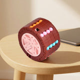 Brown cylinder fidget puzzle with a pink wheel end and colorful beads on a round wooden table beside a closed laptop.