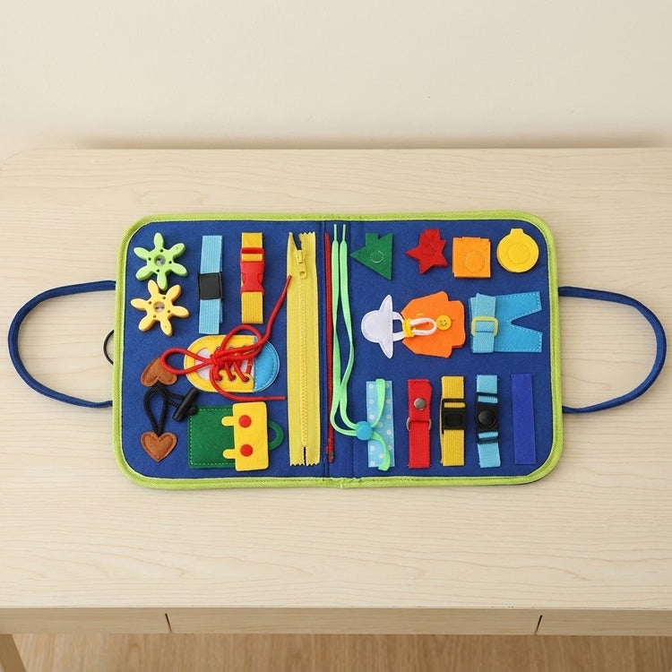 An open blue felt busy board with green edging, colorful buckles, zippers, gears and shoelaces rests on a light wooden table.