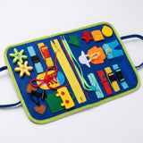 A blue felt busy board with green trim lies diagonally on white, displaying colorful fasteners, shapes, gears and a lacing shoe.
