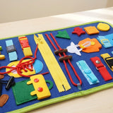 A close-up of a blue felt busy board shows a yellow zipper, red laces, colorful buckles and shapes on pale wood.