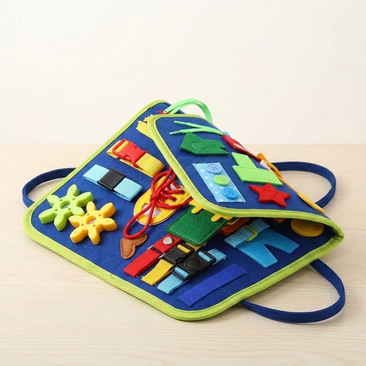 A blue felt busy board with green edging rests partly folded on pale wood, revealing layered activities and blue carry loops.