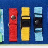 A close-up of the blue felt busy board shows red, yellow and light blue straps with a metal snap and black buckles.