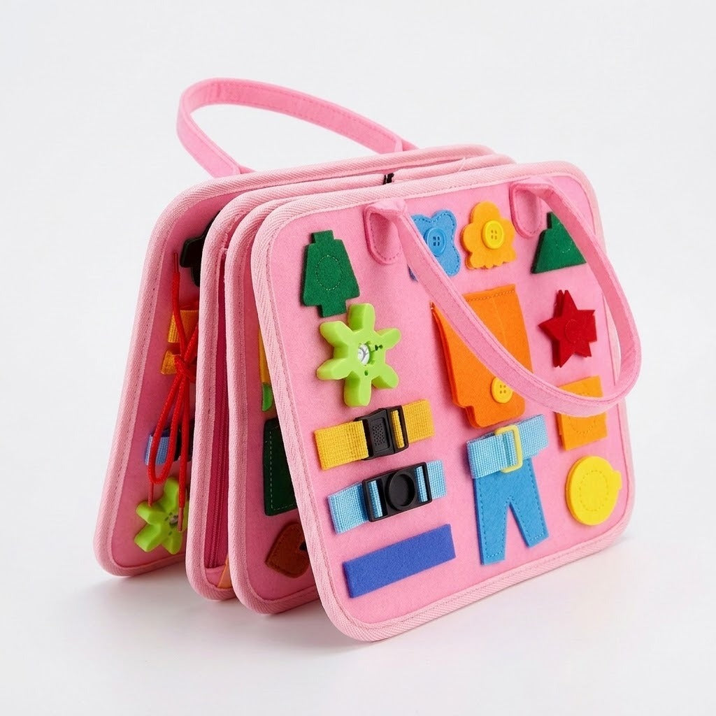A pink felt busy board stands partly open on white, showing layered pages, carry loops, colorful shapes, buckles and a green gear.