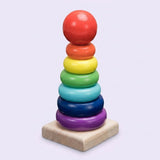 Wooden learning toy with six rainbow stacking rings and a red ball topper on a square natural wood base against a pale lavender background.