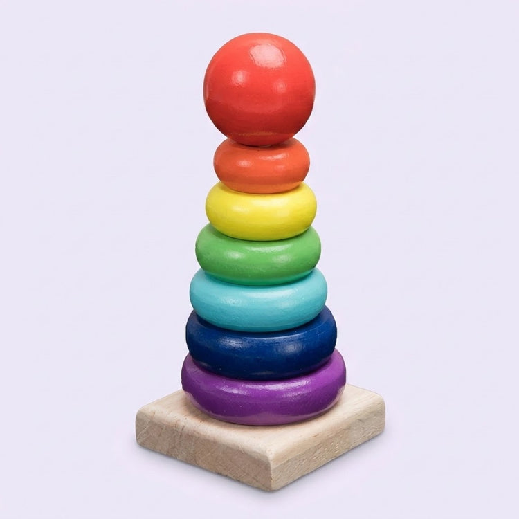 Wooden learning toy with six rainbow stacking rings and a red ball topper on a square natural wood base against a pale lavender background.
