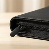 A close-up shows the black trading card binder's textured PU cover and zipper on light wood against a soft beige background.