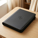 A closed black trading card binder with a textured PU cover and zipper rests on a light wooden table beside a window.