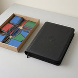 A black zippered trading card binder rests beside a cardboard tray of colorful card stacks on a light gray tabletop.