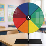 An acrylic prize wheel with eight coloured segments and a white central pointer stands on a desk in a blurred classroom.