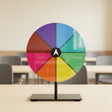 A translucent multicoloured prize wheel on a black stand sits on a light classroom table, with blurred chairs behind it.