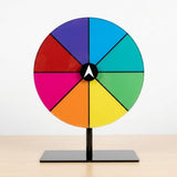 An eight-segment multicoloured prize wheel with a white central pointer stands on a light wooden surface against a plain white background.