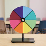 Acrylic tabletop prize wheel with eight multicolored segments on a black stand, on a light wood table in a blurred classroom.