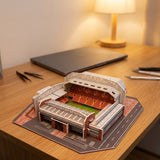 A plastic 3D stadium puzzle with red seating and white roof supports sits on a wooden desk beside a closed laptop.