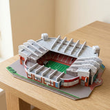 A plastic 3D stadium puzzle with gray roofing, red seating and a green pitch sits on a light wooden table.