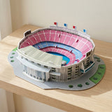 A plastic 3D stadium puzzle with pink and blue seating sits on a wooden shelf against a cream wall.