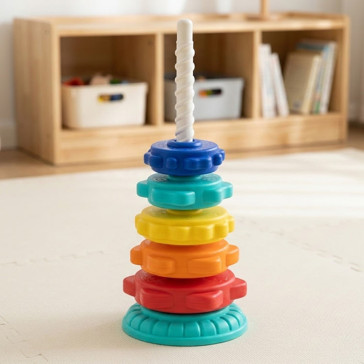 A colorful plastic stacking ring toy with a white spiral pole stands on a light-colored floor in front of a wooden storage shelf.