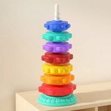 A stacking toy with seven colourful plastic rings, a white spiral post and mint base stands on a shelf against a cream wall.