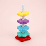 A stacking toy with yellow, purple, blue and turquoise flower-shaped rings, a white spiral post and red base stands against a pink background.