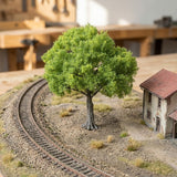 A light green miniature tree stands beside curved model railway tracks and a small house on a textured landscape.
