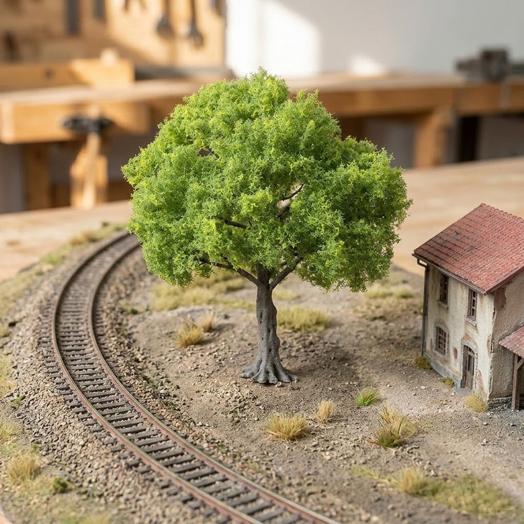 A light green miniature tree stands beside curved model railway tracks and a small house on a textured landscape.