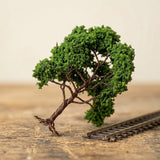 A green miniature tree with exposed brown twisted wire branches leans beside a short model railway track on a workbench.