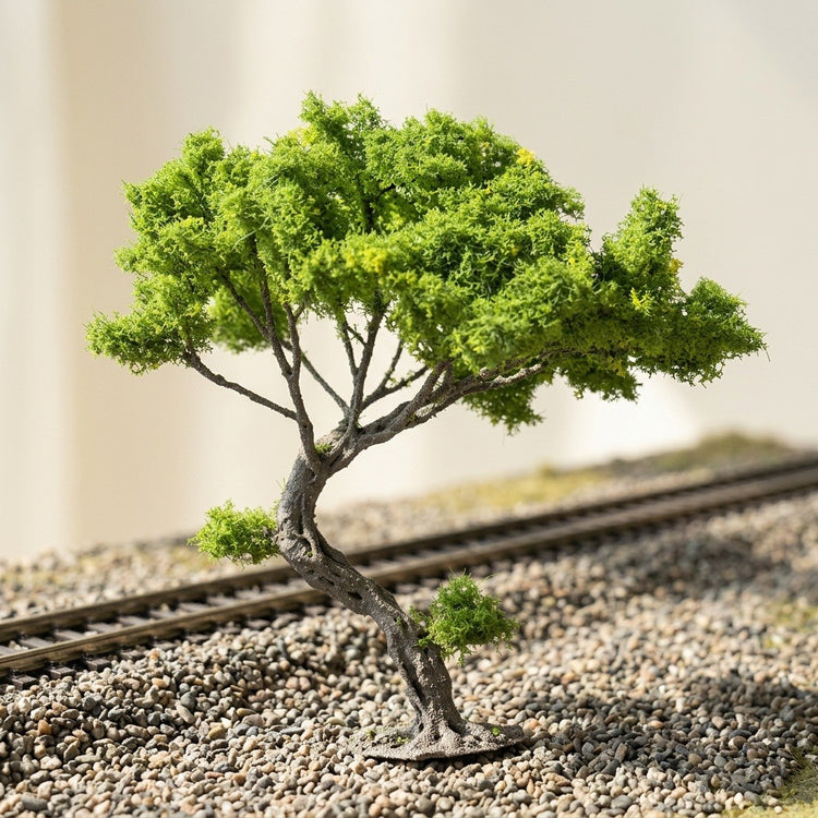 A miniature tree with green foliage and a curved gray trunk stands on gravel beside model railway tracks.
