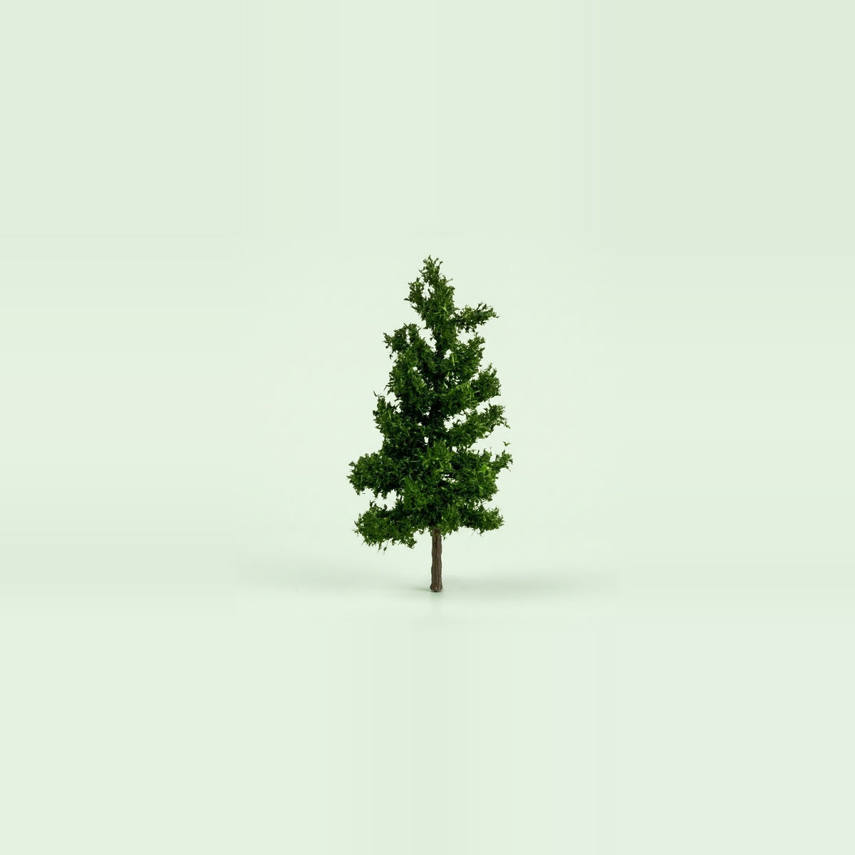 A dark green miniature model pine tree with a slim brown trunk on a pale green studio background, 3.9 inches (10 cm) tall.